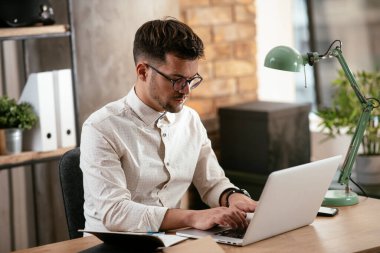 Gözlüklü ve beyaz gömlekli genç adam çalışma masasında dizüstü bilgisayarlı.