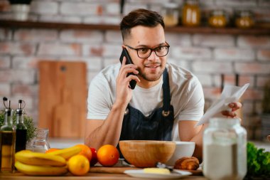 Önlük giyen genç adam mutfakta telefonla konuşuyor.