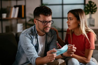 Sağlık maskeli genç bir adam ve kız arkadaşı oturma odasında oturuyor.