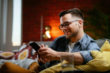 Genç adam kanepede uzanıp müzik dinliyor. Modern adam oturma odasında telefonuyla müzik dinliyor..
