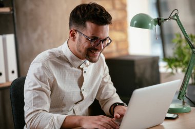 Gözlüklü ve beyaz gömlekli genç adam çalışma masasında dizüstü bilgisayarlı.