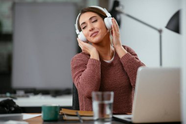 Ofiste kulaklıklı genç bir kadın. Genç gündelik iş kadını en sevdiği şarkıyı kulaklıkla dinliyor ve ofiste dizüstü bilgisayarla çalışırken şarkı söylüyor..