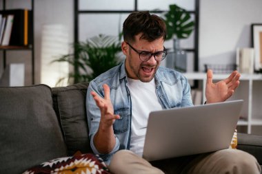 Oturma odasında dizüstü bilgisayarı olan stresli genç bir adam.