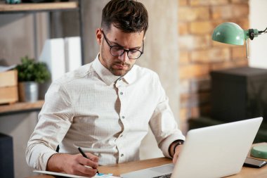 Gözlüklü ve beyaz gömlekli genç adam çalışma masasında dizüstü bilgisayarlı.