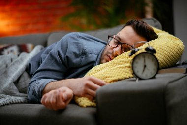 Genç adam çalar saatle kanepede yatıyor.