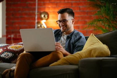 Oturma odasında dizüstü bilgisayarlı gözlüklü gülümseyen genç adam.