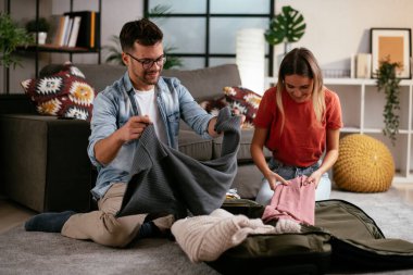 Genç adam ve kız arkadaşı oturma odasında bavul hazırlıyorlar.