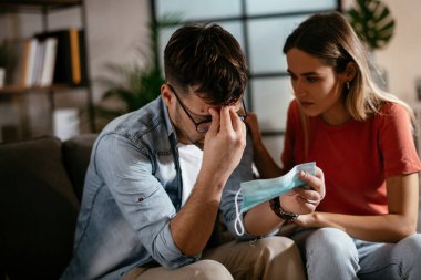 Sağlık maskeli genç bir adam ve kız arkadaşı oturma odasında oturuyor.