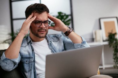 Oturma odasında dizüstü bilgisayarı olan stresli genç bir adam.