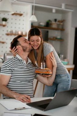Masada çalışan bir adam, mutfakta ona kek sunan bir kadın.