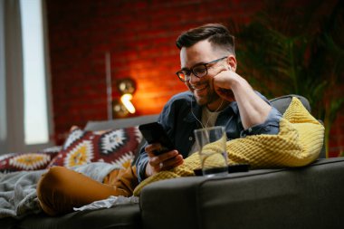 Genç adam kanepede uzanıp müzik dinliyor. Modern adam oturma odasında telefonuyla müzik dinliyor..