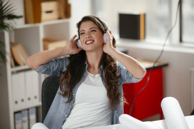 Ofiste kulaklıklı genç bir kadın. Genç gündelik iş kadını en sevdiği şarkıyı kulaklıkla dinliyor ve ofiste dizüstü bilgisayarla çalışırken şarkı söylüyor..