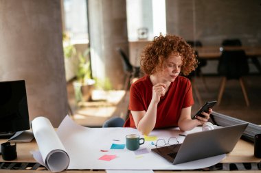 Güzel bir iş kadını proje üzerinde çalışıyor. Ofiste akıllı telefon kullanan kıvırcık saçlı genç bir kadın..