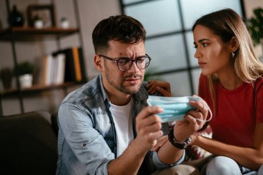 Sağlık maskeli genç bir adam ve kız arkadaşı oturma odasında oturuyor.