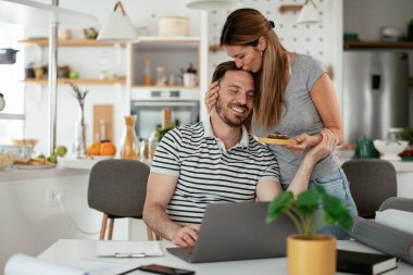 Masada çalışan bir adam, mutfakta ona kek sunan bir kadın.