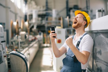 Sarı miğferli mutlu işçi fabrikada telefon kullanıyor. 