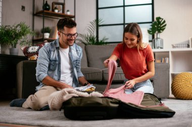 Genç adam ve kız arkadaşı oturma odasında bavul hazırlıyorlar.