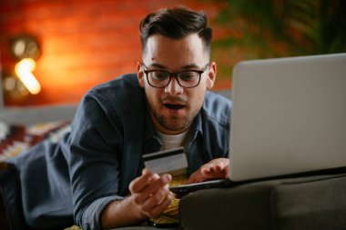 Genç adam oturma odasında online alışveriş için kredi kartı kullanıyor..