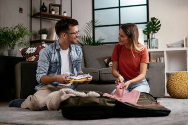 Genç adam ve kız arkadaşı oturma odasında bavul hazırlıyorlar.