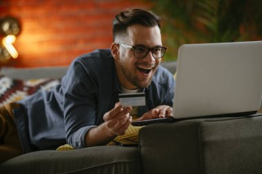 Genç adam oturma odasında online alışveriş için kredi kartı kullanıyor..