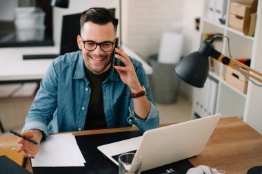 Genç adam çalışma masasında telefonla konuşuyor.