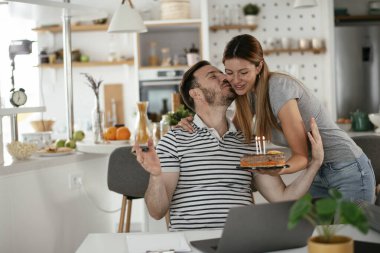 Masada çalışan bir adam, mutfakta ona kek sunan bir kadın.