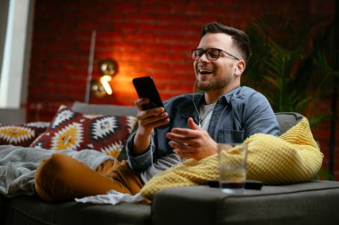 Genç adam kanepede uzanıp müzik dinliyor. Modern adam oturma odasında telefonuyla müzik dinliyor..