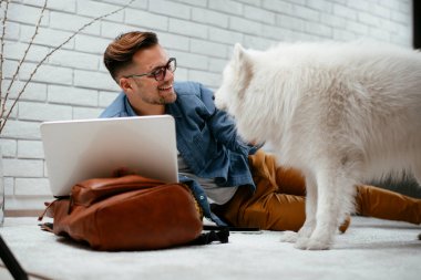Adam dizüstü bilgisayarla çalışırken köpeğiyle yerde oynuyor. Samoe köpeğiyle oynayan adam..