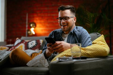 Genç adam kanepede uzanıp müzik dinliyor. Modern adam oturma odasında telefonuyla müzik dinliyor..