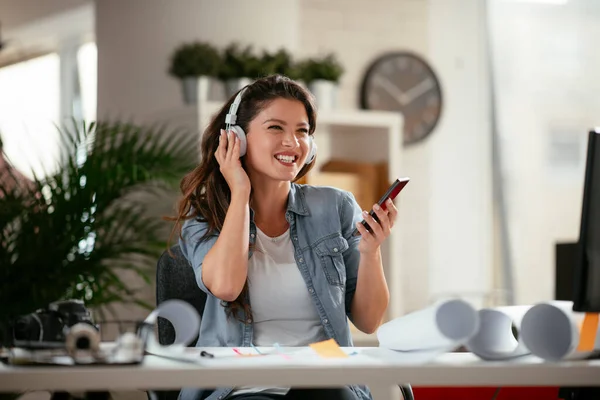 Young woman in office with headphones. Young casual businesswoman enjoying in her favorite song over headphones and singing while working on laptop in the office.