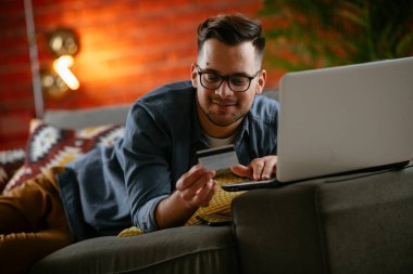 Genç adam oturma odasında online alışveriş için kredi kartı kullanıyor..