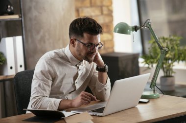 Gözlüklü ve beyaz gömlekli genç adam çalışma masasında dizüstü bilgisayarlı.