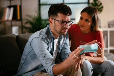 Sağlık maskeli genç bir adam ve kız arkadaşı oturma odasında oturuyor.