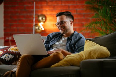 Oturma odasında dizüstü bilgisayarlı gözlüklü gülümseyen genç adam.