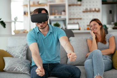 Mutlu çift oturma odasında sanal gerçeklik gözlükleriyle eğleniyor. Evde video oyunu oynayan ve eğlenen genç bir çift..