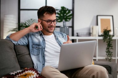 Oturma odasında dizüstü bilgisayarlı gözlüklü genç adam.