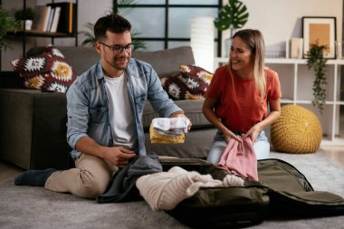 Genç adam ve kız arkadaşı oturma odasında bavul hazırlıyorlar.