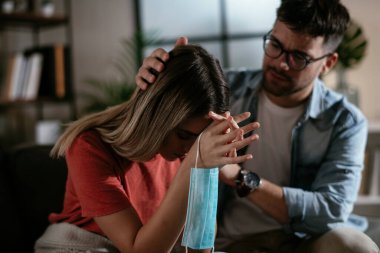 Tıbbi maskeli genç bir kadın ve erkek arkadaşı oturma odasında oturuyor.