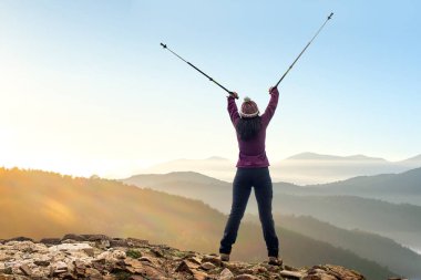 Genç bayan yürüyüşçünün havada sırık kaldırışının arka görüntüsü. Dağın tepesinde kayaların üzerinde duran kadın..