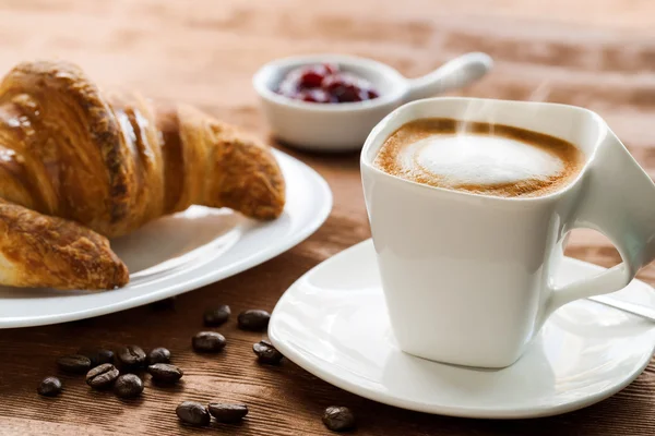 Creamy cup of coffee with croissant in background. — Stock Photo, Image