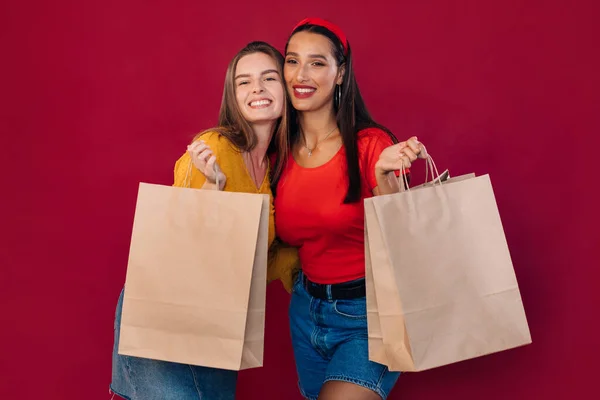 两个穿着休闲装的年轻的黑发女友在购物后拿着购物袋摆姿势 与红色背景隔离 — 图库照片