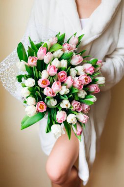 Beyaz sabahlık giymiş, pembe ve beyaz lalelerle poz veren genç ve güzel bir kadın..