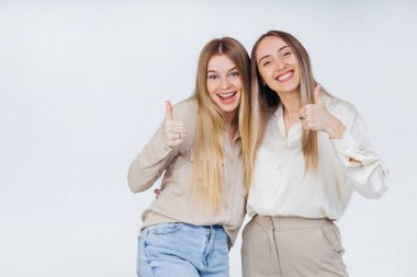 Gri arkaplanda el hareketi olarak baş parmak gösteren tişörtlü iki neşeli genç bayan arkadaş..