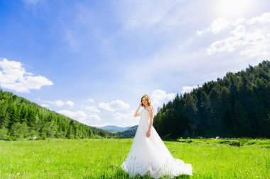 Esmer, güzel gelin kız, dağların arka planında beyaz bir gelinlikle tarlada yürüyor..