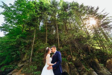 Romantik evli çift. Gelin ve damat birbirlerine yaslanırlar. Kafasında çelenk olan bir gelin. Uzun saçlı genç gelin. Dağın arka planına karşı.