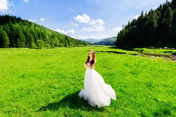 一位漂亮的卷发新娘穿着一条长长的花边白色连衣裙 站在草地上的草地上 背靠着青翠的植物和树木 — 图库照片