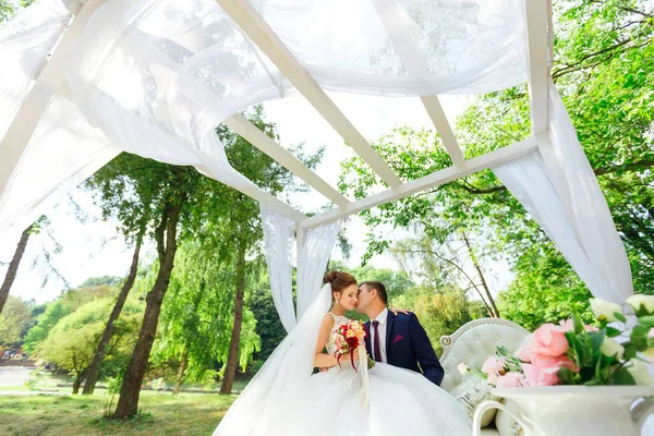 花嫁と新郎は白いベンチに座って 空飛ぶ布で作られたアーチの下に抱きついています — ストック写真