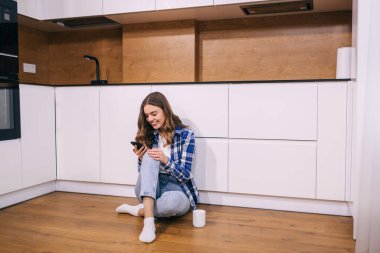 Bir fincan çayla tişörtlü güzel genç bir kadın yerde oturmuş, sosyal ağları kontrol ediyor..