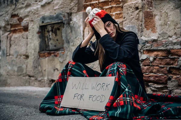 A homeless woman sits on the street in the shade of a house and asks for help and money. Problems of large modern cities. Grasp your head with your hands. Poverty, drugs.