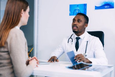 Ciddi bir Afrikalı Amerikalı doktor genç bir hastayla konuşuyor ve ofisinde otururken notlar alıyor.